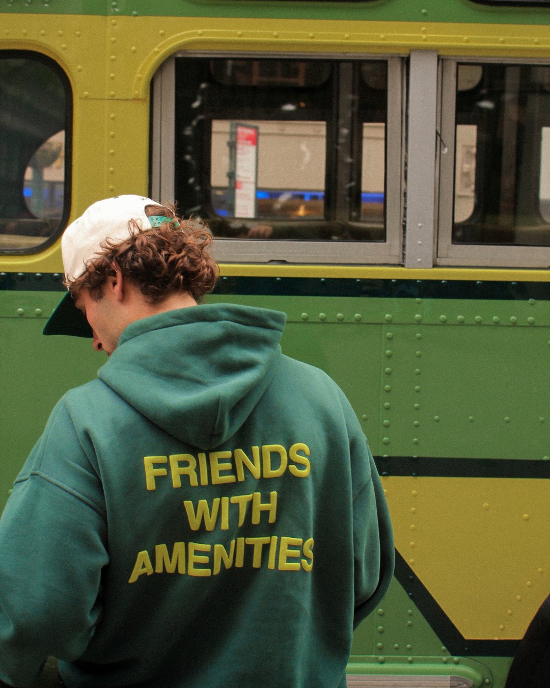 A traveler in a Friends With Amenities hoodie beside a vintage green bus.
